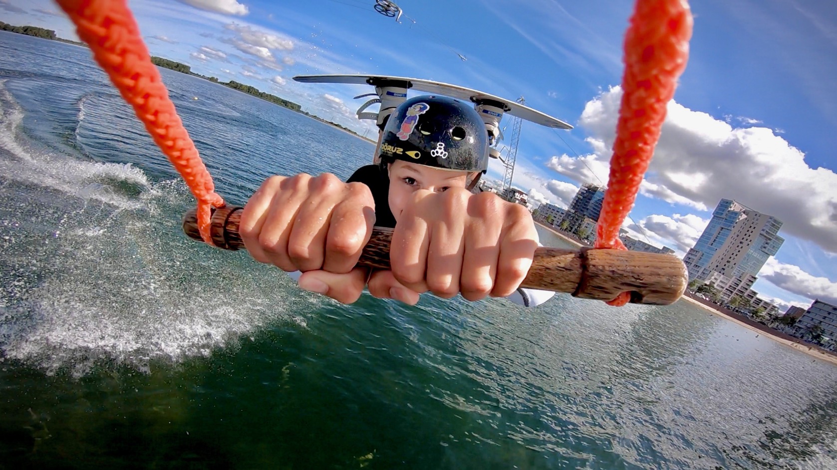 Wakeboarder gripping the cable tow handle with the tower cable and Rotterdam skyline visible behind